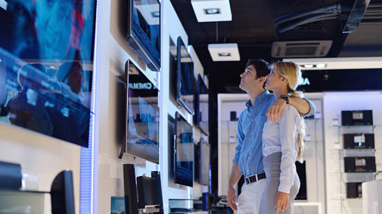 Young couple in consumer electronics store looking at latest laptop, television and photo camera to buy lcd flat tv